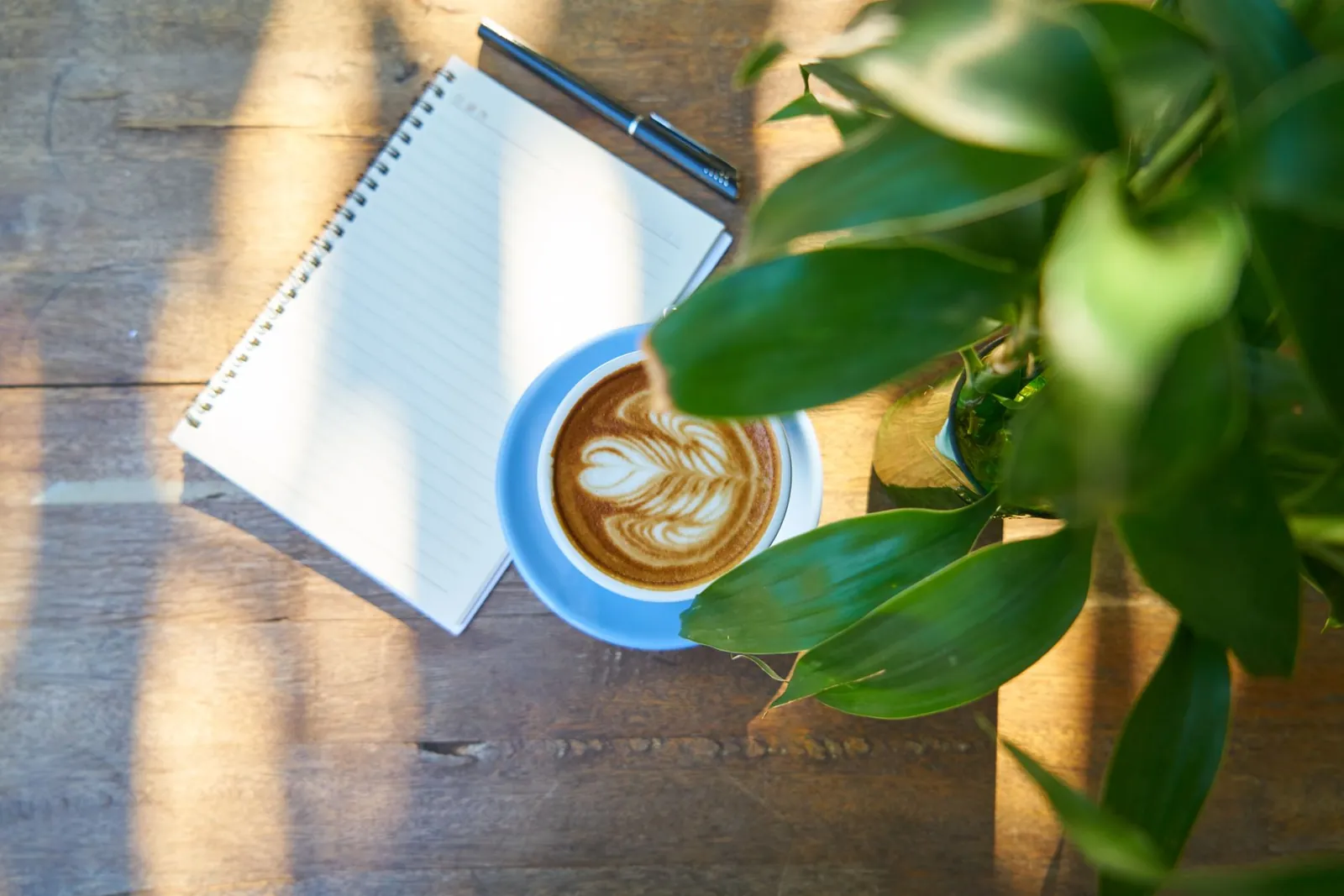 An overhead view of a notebook, pen and coffee on a warm wooden desk
